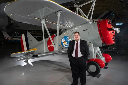 A white man in his 30s stands in front of a restored biplane displayed on a polished concrete floor inside a museum. He is wearing glasses and a dark suit with a white shirt and burgundy tie.