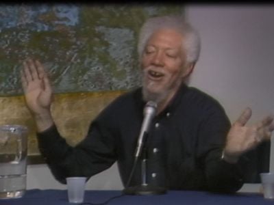 Benny Andrews sits at a table with a microphone, cups, and a pitcher of water with a yellow, green, and blue abstract painting hanging behind him. He is wearing a black button down shirt and raising his hands while talking.