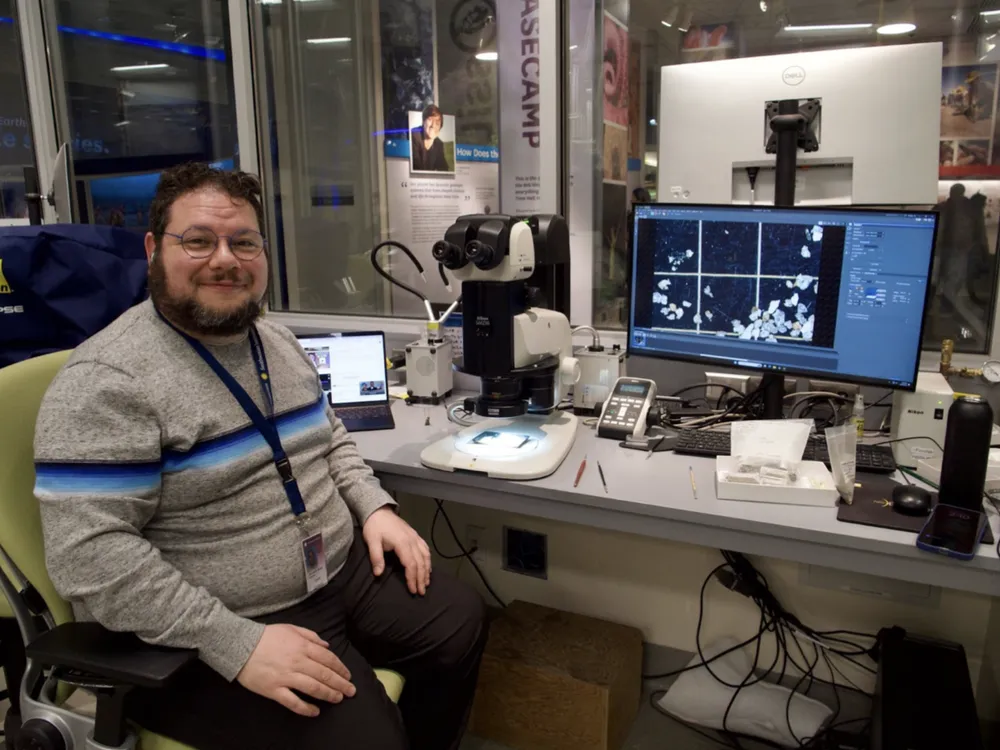A man in a grey short sits in a lab setting, in front of a microscope and a computer screen.