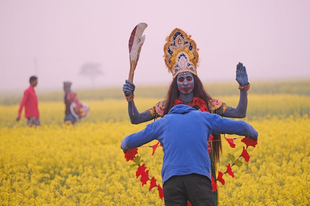 A folk artist known as Bohurupee is getting his garland adjusted by a passing villager as he is on his walk through flowering mustard fields.