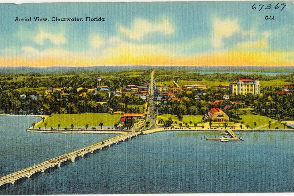 An aerial view of the city on the bay, with blue water in foreground and a bridge extending to the left