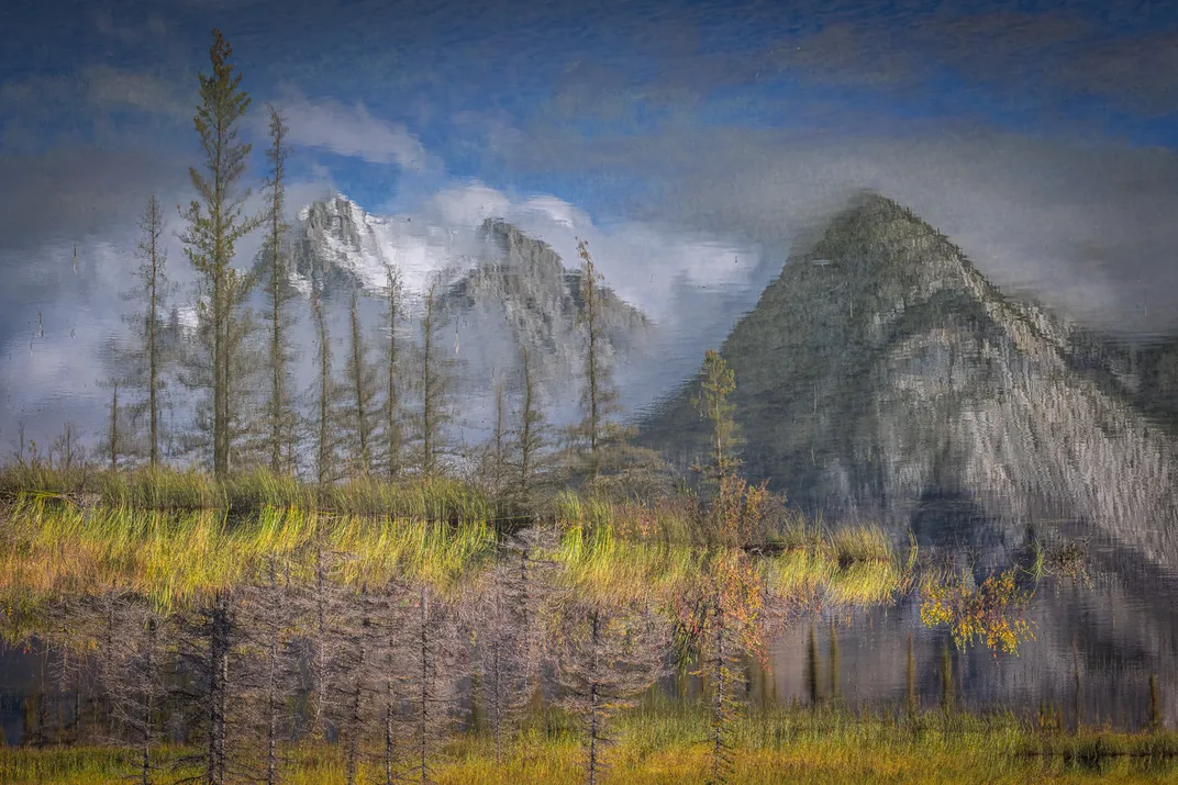 a landscape of mountains, sparse trees and yellow grasses is reflected in a lake surface, with the real scenery seen flipped upside-down at the bottom