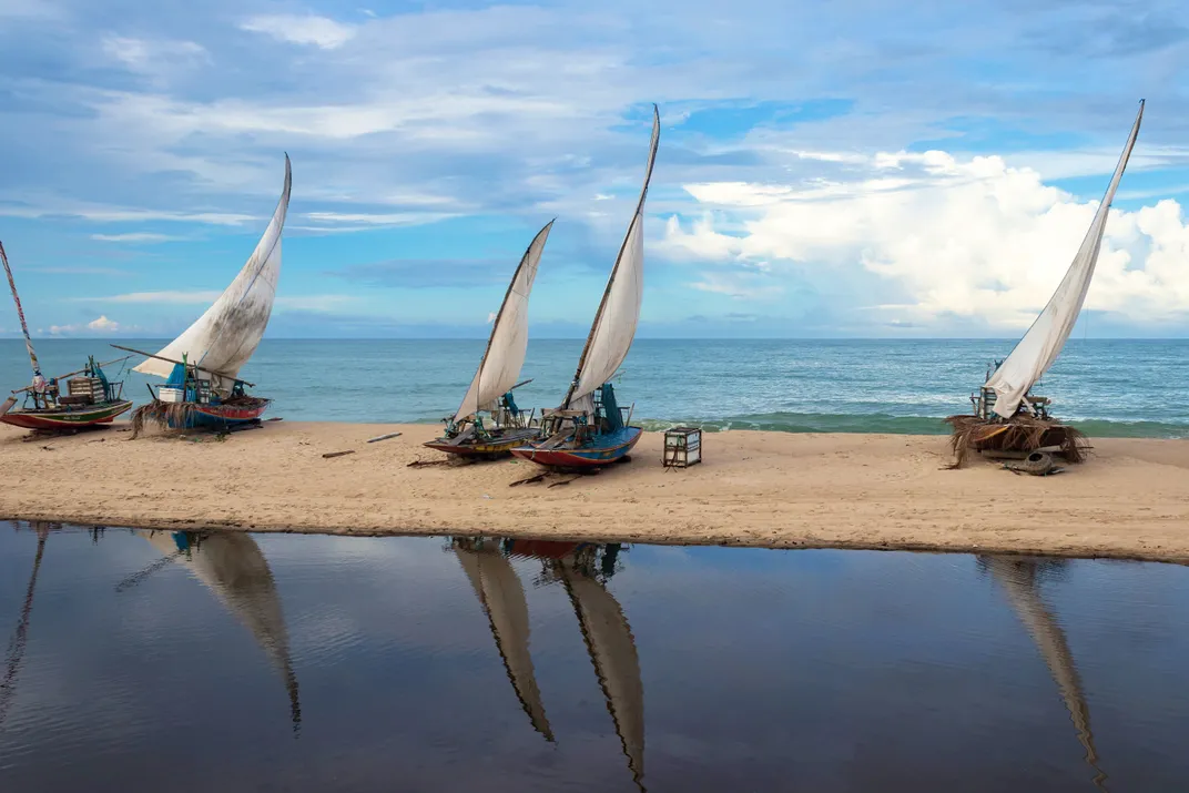 The tall sails of boats are reflected in the waters next to a sandbar off a Brazilian beach.