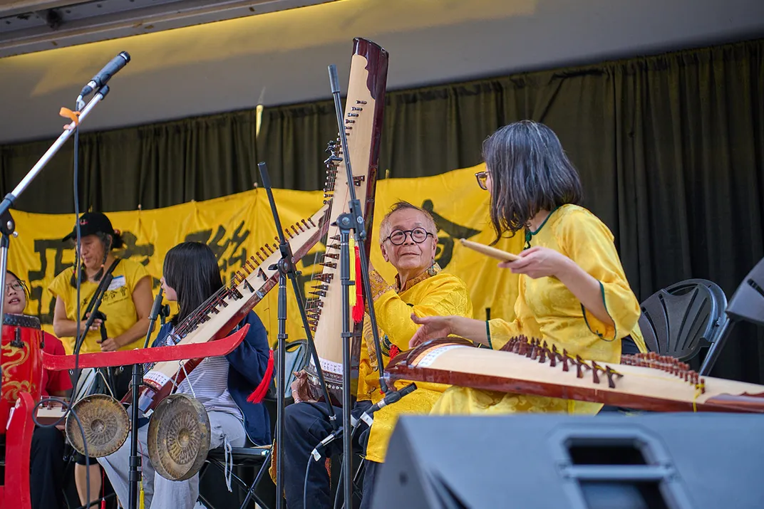 Musicians prepare to play on stage, holding long stringed zither instruments. A pair of small metal gongs hangs in front of them.
