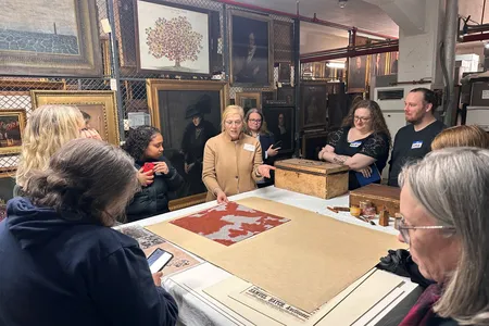 A group of workshop participants standing around a table listening to a curator discuss an artifact within Historic New England’s collection.