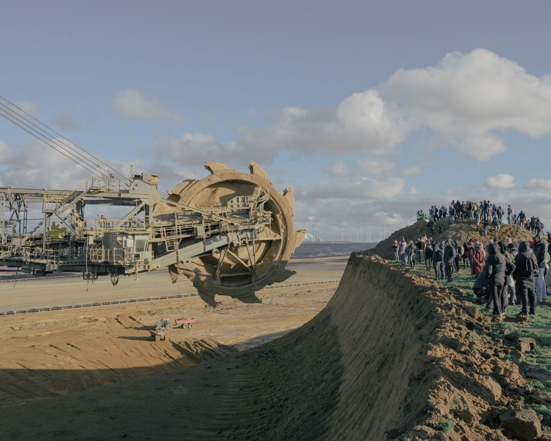 people stand on a cliff staring down a massive construction toothed wheel