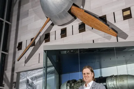 A white man in his late 30s stands inside a gallery displaying aviation artifacts, including a four-blade fan mounted on the wall.