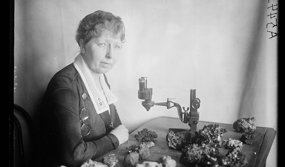 Black and white photo of Margaret W. Moodey sitting at a desk with a microscope and gemstones