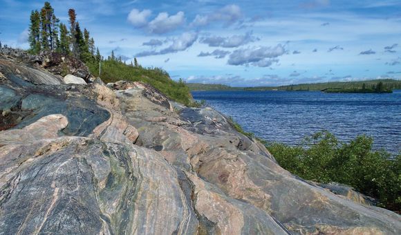 A slab of multicolored, sedimentary rock is pictured with vibrant trees and water surrounding it.