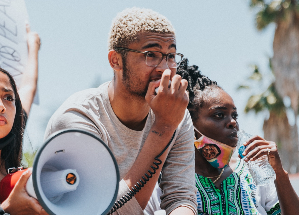 Three young activists are outside at a protest – looking off into the distance. They all appear to be African American. One is holding a bullhorn and has a determined look on her face. Another is holding the bullhorn’s microphone and speaking into it.