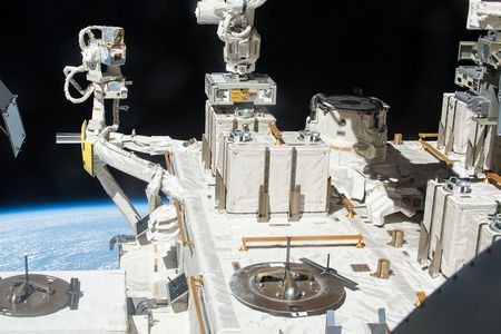 A robotic arm places a container with three panels of bacteria outside the International Space Station.