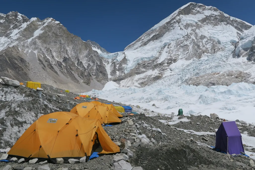 A photo of Everest Base Camp. Tents of various colors are scattered across the gray, rocky ground. Snow-covered mountains are in the background.