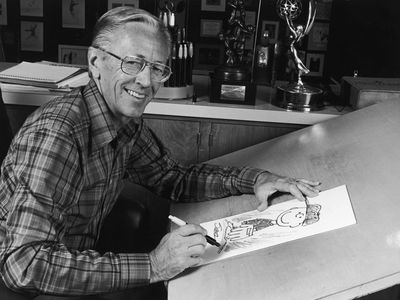 Charles M. Schulz, creator of the &quot;Peanuts&quot; comic strip, at his studio drawing table with a picture of his character Charlie Brown and some awards behind him in 1978