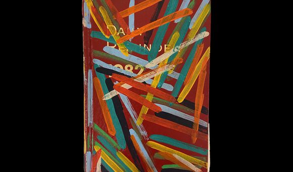 A photograph of a red book that reads, “Daily Reminders 1982” in white lettering. The book has yellow, green, blue, red, and orange strips of paint on it.