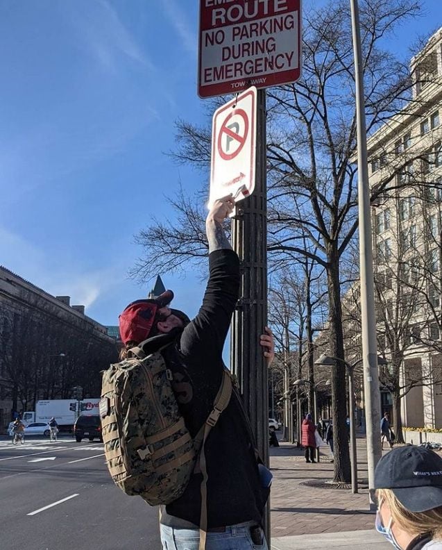 A person wearing a camouflage backpack leans against a street-pole, using a wedged tool to remove stickers from a "No Parking" sign.