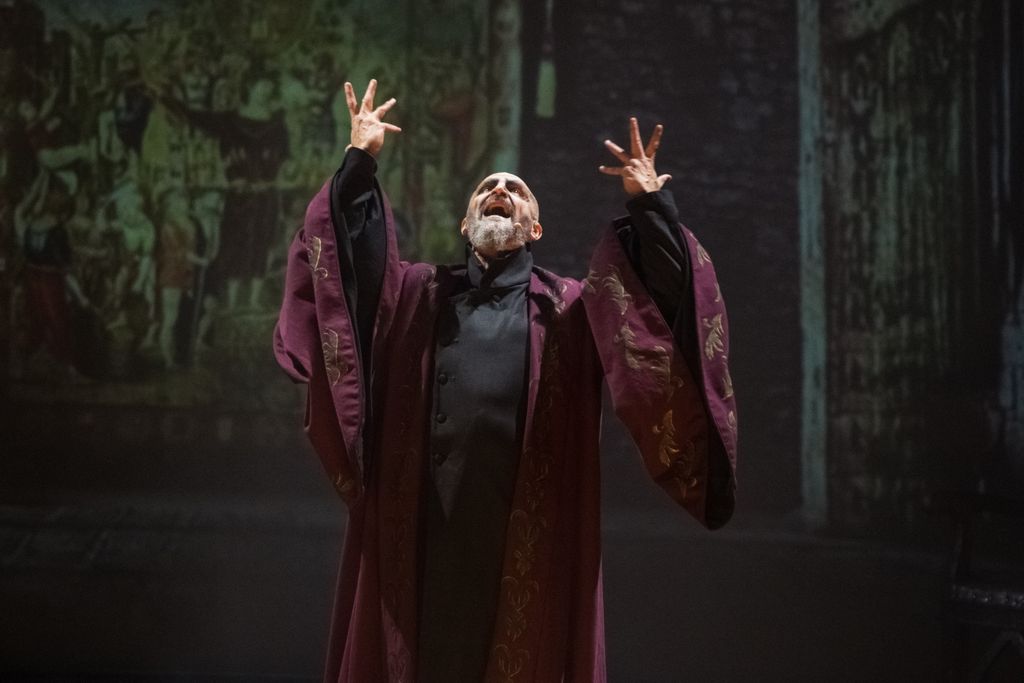 Man wearing a burgundy robe sings with arms outstretched on stage
