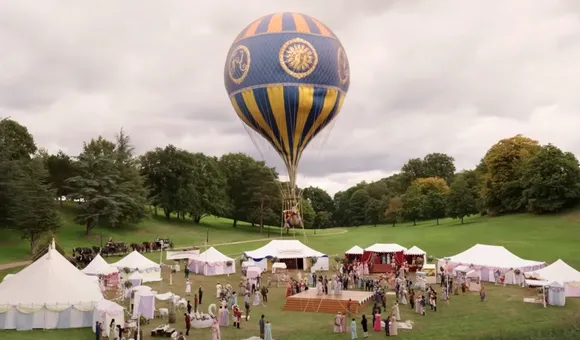 A vibrant hydrogen balloon with intricate blue and gold designs floats above a bustling outdoor event set against a cloudy sky. Below, several white tents are set up in a grassy park, with groups of people mingling and walking around.