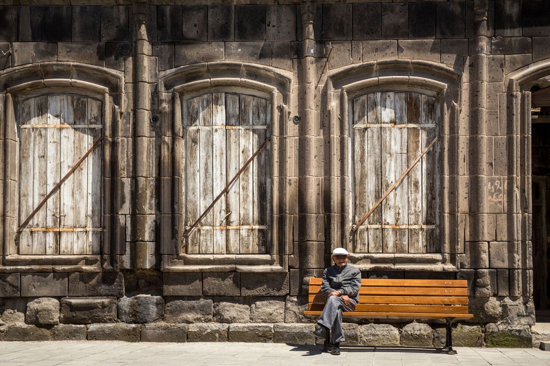 A man sits in front of a dark grey, stone wall with three stately wooden doors.