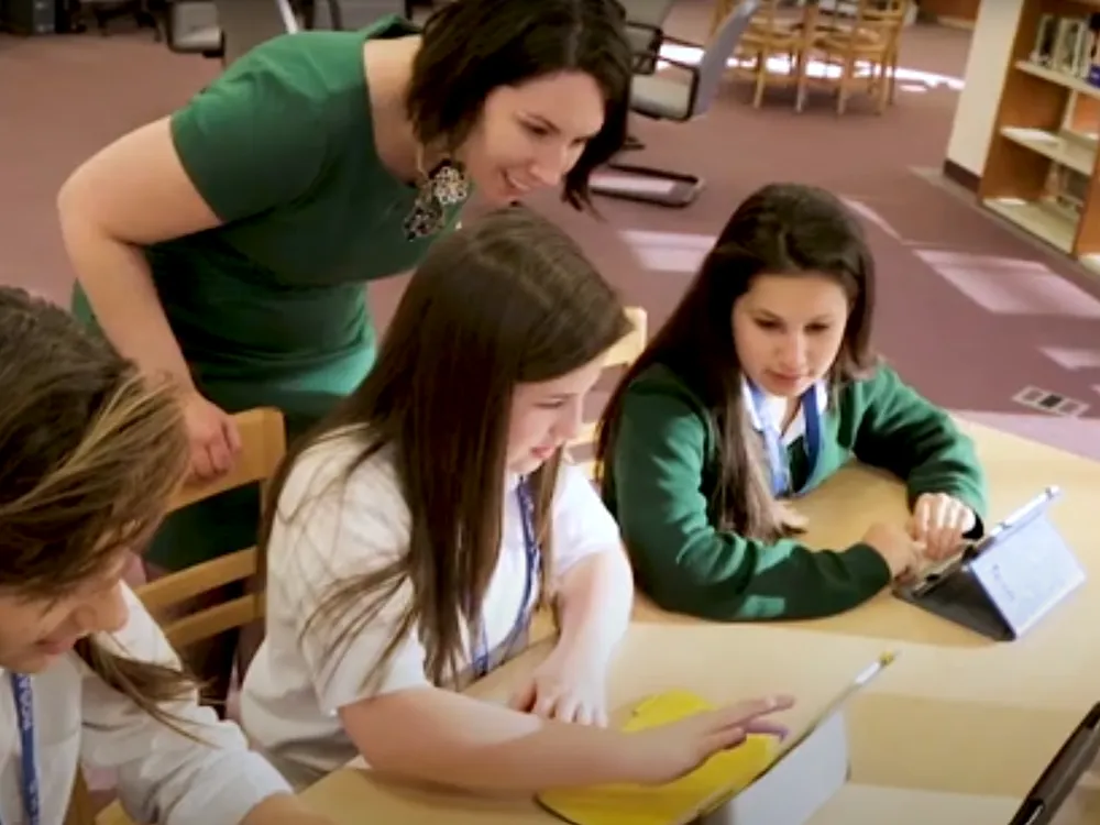 Students are working on their laptops and tablets while a teacher looks over their shoulders