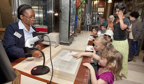 A group of students wait in line at a Post Office. Each student has a stamped envelope they are handing to the postal worker behind the counter.