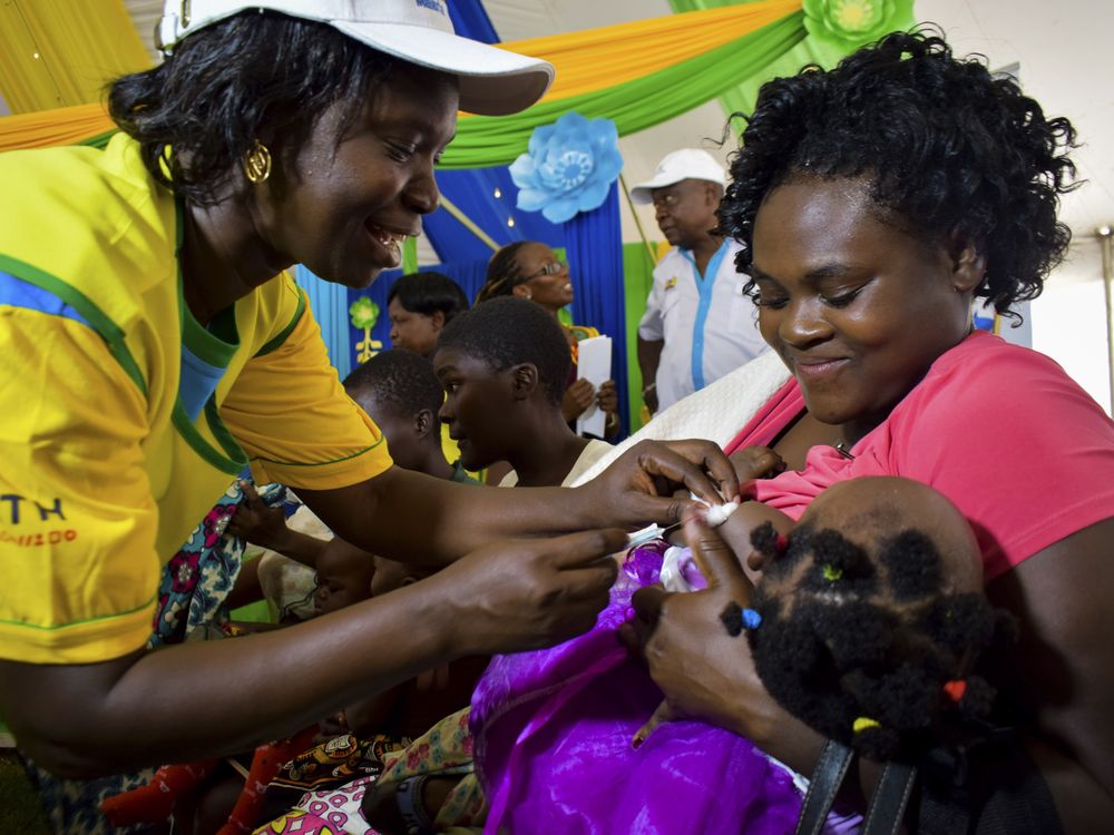 A health worker vaccinating a child against malaria in Kenya. The child is being held by her mother who is smiling.