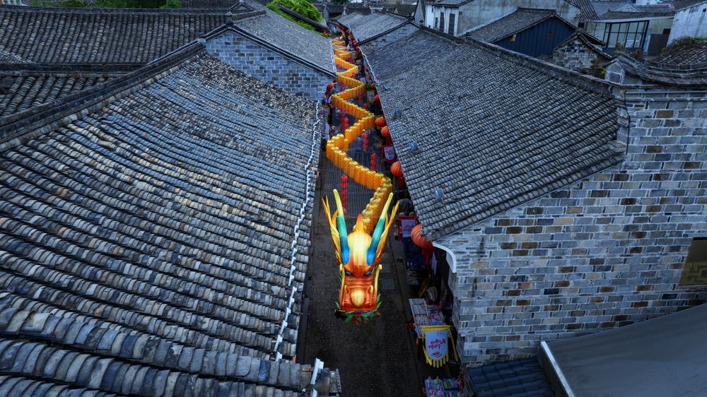 Shot from above with a drone, an ancient alley unfolds between rows of gray-tiled rooftops as a vividly colored dragon winds its way through the historic architecture. The bright dragon contrasts strikingly with the textured surfaces of the old buildings, creating a lively and festive scene rich in traditional folk culture.