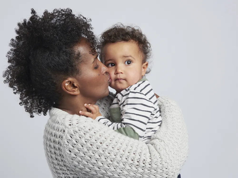 A mom holds her toddler and gives him a kiss on the cheek against a grey background
