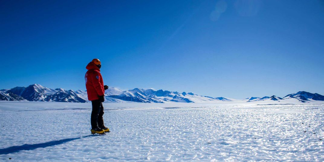 A scientist in a red coat and orange helmet stands on the sunny Antarctic ice sheet, with blue mountains stretching in the background