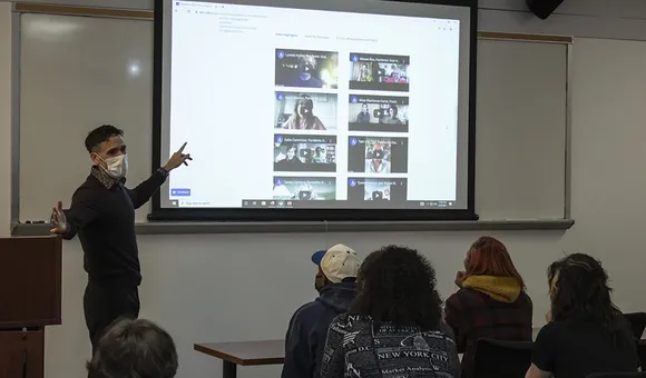 A man points to a video screen in a classroom with students seated in the foreground.