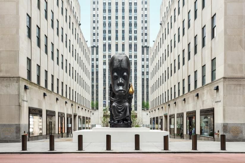 A view of a courtyard surrounded by tall white buildings with windows on all sides; in the middle, a large sculpture that resembles a man with a hugely oversized head sitting on a throne, cast in all black bronze