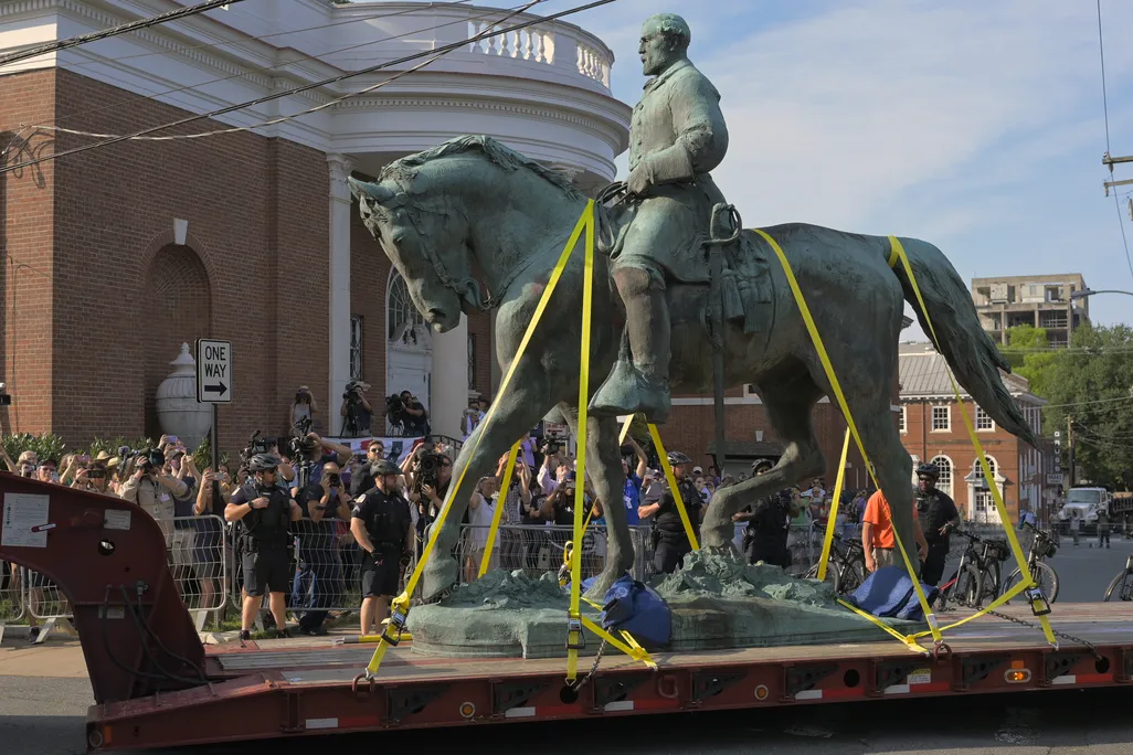 Construction crews transport a greenish bronze statue of Lee on horseback past a crowd of onlookers on a truck bed