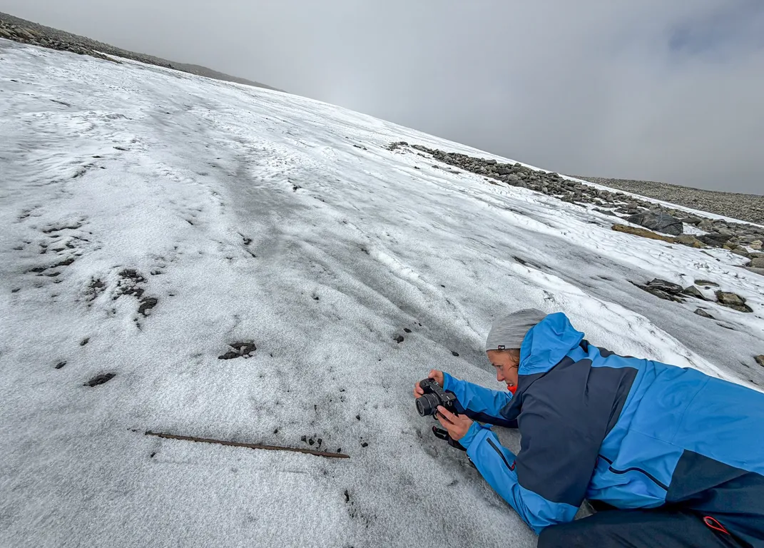 A person in a blue coat taking a photo of an artifact on some ice