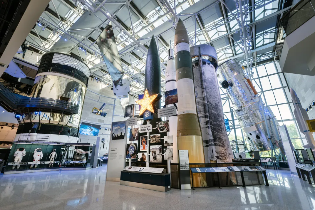 various rockets and the cylindrical Skylab are on display in an atrium with windows in the ceiling and back wall. A sign reads "RTX Living in the Space Age"