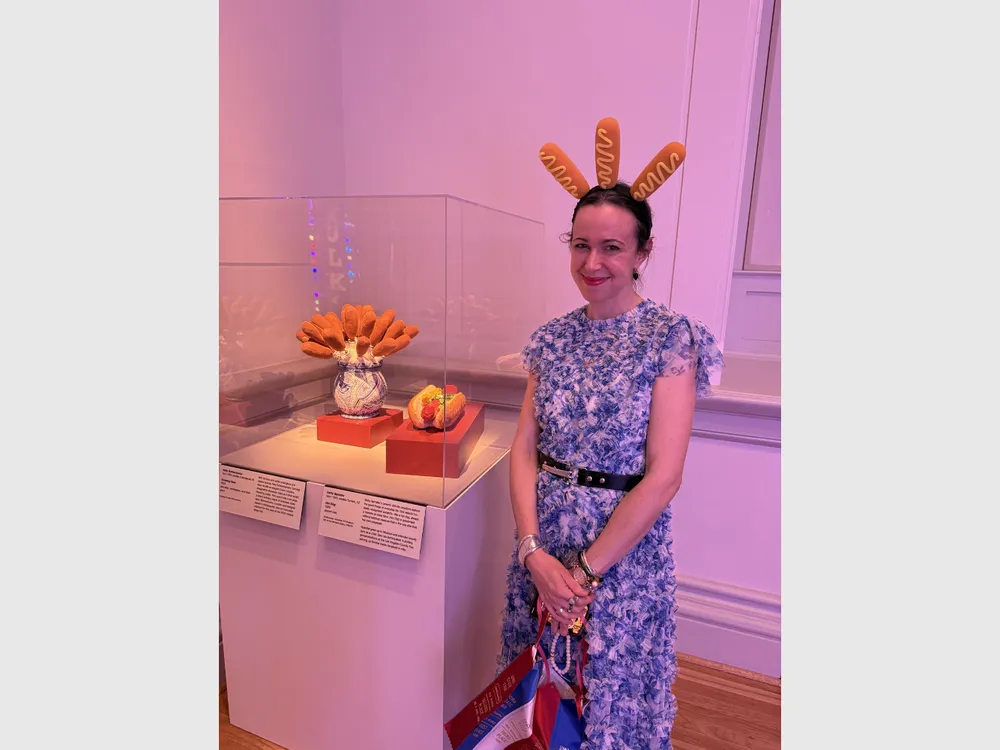 Sarah Rogers Morris wearing a headband with three corndogs poking out stands behind an exhibition case with a vase containing corndogs and a ceramic hot dog.
