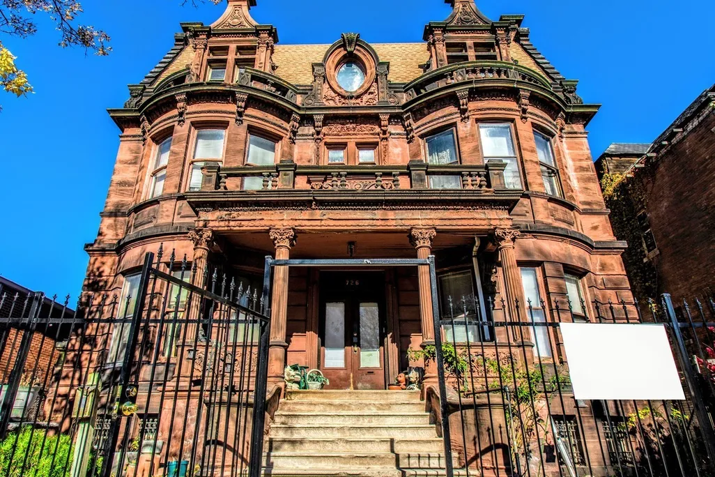 A front view of an ornate brownstone mansion, with columns, a circular window in the center, turret-like shapes on either side and a peaked roof