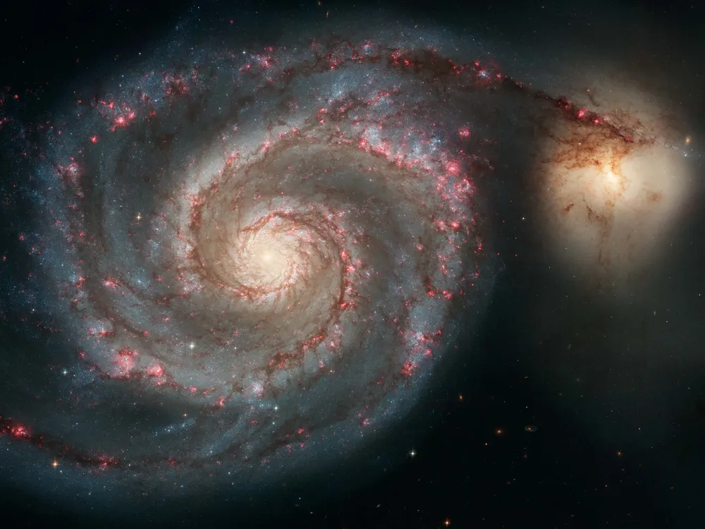 a swirling spiral galaxy with a yellow center and arms with blue and pink, one arm stretches and interacts with another galaxy