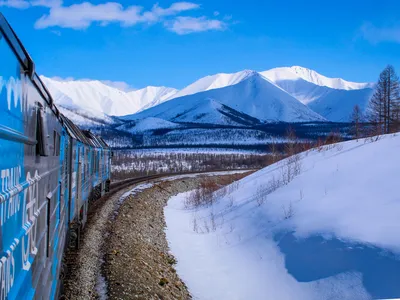 A train whizzes through snowy hills with frosty peaks from the Stanovoy Ridge mountains in the distance.