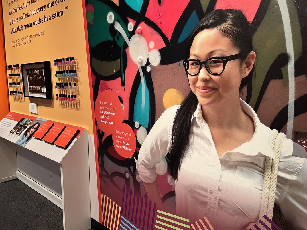 A large wall-sized photo mural of Tina Pham in prominently visible in a museum exhibit. Tina is a Vietnamese-American woman with black glasses. She is wearing a white button-down shirt and has her hands on her hips. The photo is taken from the waist up.