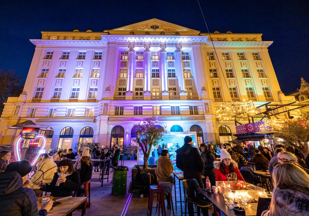 The Esplanade Zagreb Hotel illuminated with colorful holiday lights at night, overlooking a busy Advent Zagreb market with food stalls, outdoor seating, and festive decorations.
