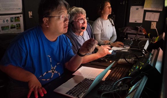 Onboard science leads Drs. Chris Mah and Chris Kelley and NOAA Educational Partnership Program Intern Nikola Rodriguez discuss and take a closer look at deepwater habitats explored with remotely operated vehicle Deep Discoverer on Horizon Guyot. (Image courtesy of the NOAA Office of Ocean Exploration and Research, 2017 Laulima O Ka Moana)