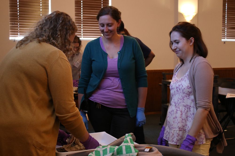 Two caucasian women stand across a table from a woman holding wet cloth.