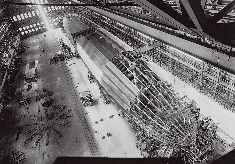 A black and white photo of an airship under construction, taken from above. Half the airship is covered with silver-colored cloth while the other half reveals internal struts and girders.
