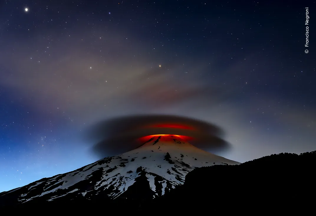a volcano erupts lava at night, beneath a double lenticular cloud, which looks like two stacked dinner plates