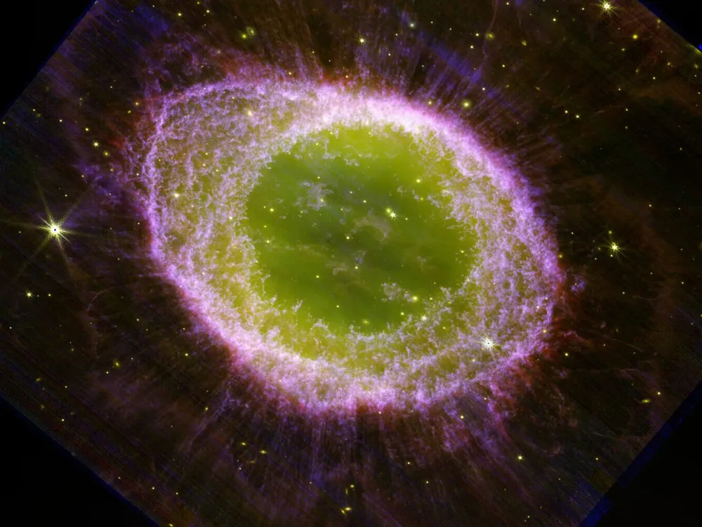 An image of the ring nebula, with a large green center, a purple outer ring, and wisps of purple beyond that. Stars and other galaxies glow yellow in the background.