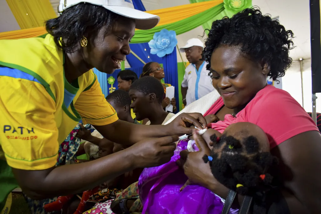 A health worker vaccinating a child against malaria in Kenya. The child is being held by her mother who is smiling.