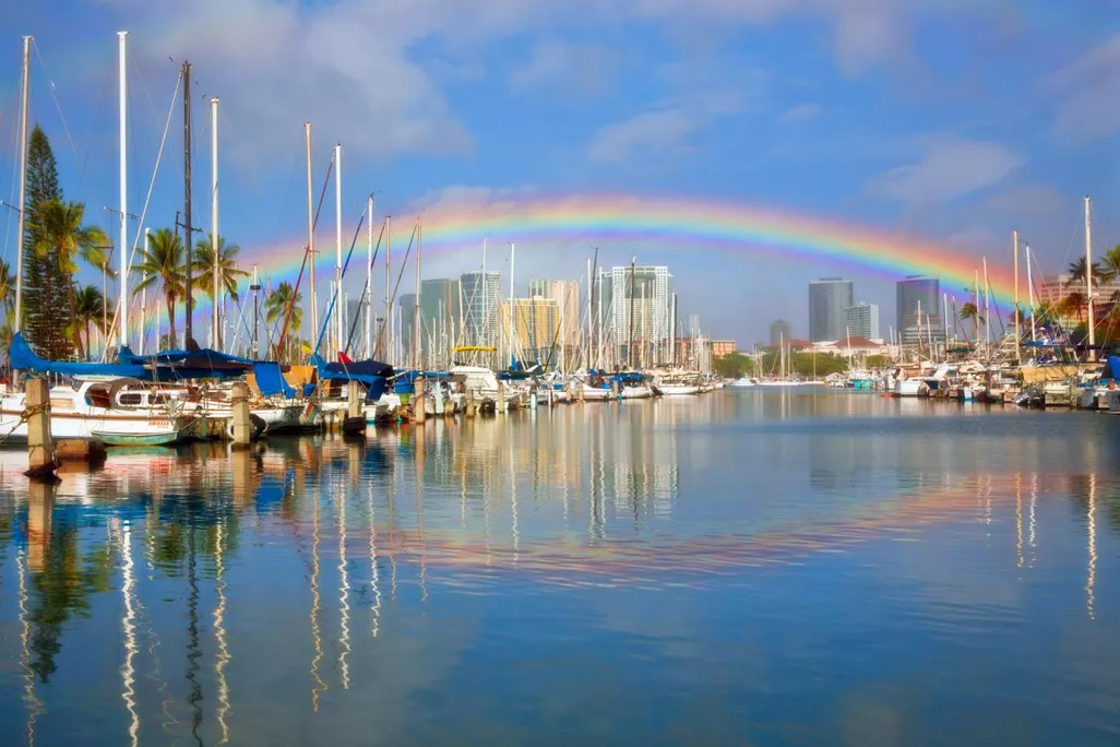 A photo of a rainbow stretching across Honolulu harbor. The photo shows various boats in the water and and a reflection of the rainbow in the water