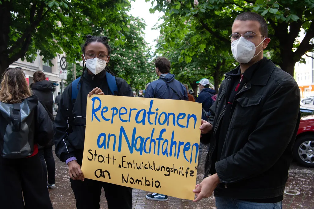 Two people in masks hold a yellow sign with blue and black handpainted letters, standing in a city street in front of trees and other protesters
