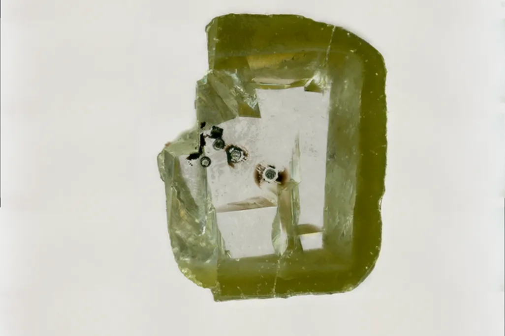 A close-up photo of davemaoite. The diamond looks like a chipped, green square. Inside, it has a few black dots that are the davemaoite.