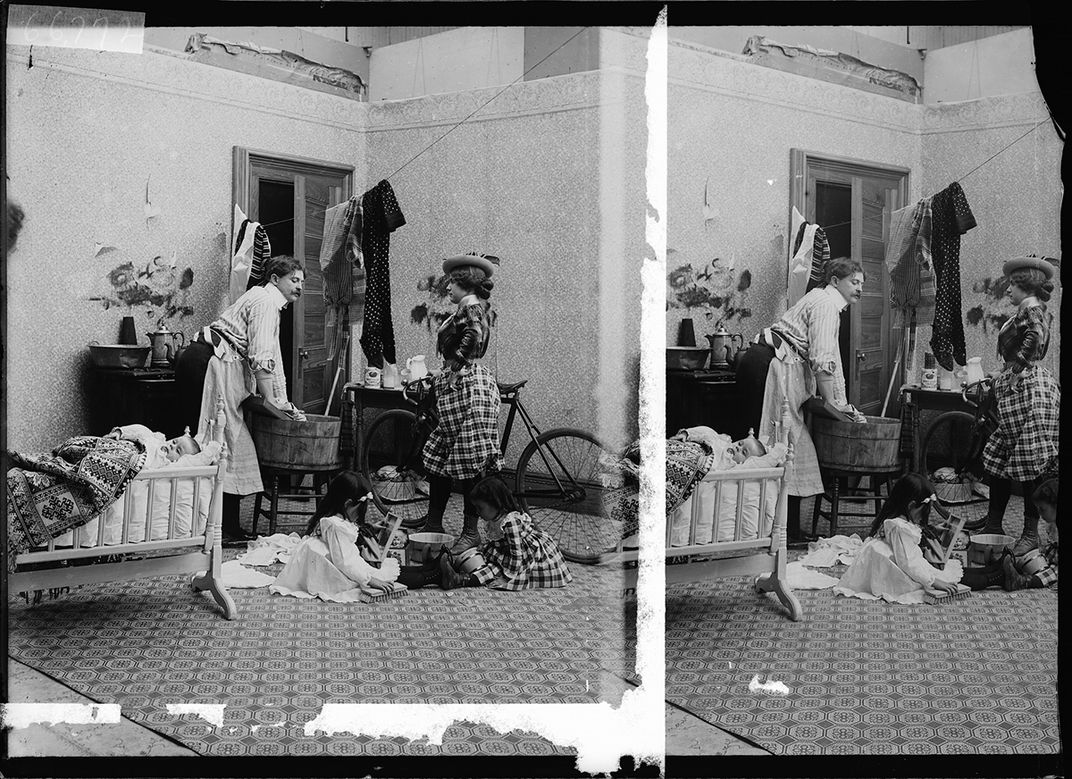 A black and white photograph shows a man doing domestic labor while a woman prepares to go out on a bicycle.