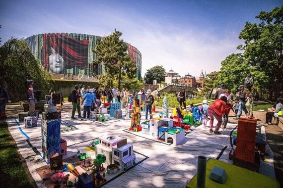 A large outdoor space is filled with human-sized colorful cardboard structures. In the background, the Hirshhorn Museum and Smithsonian Arts and Industries building are visible.